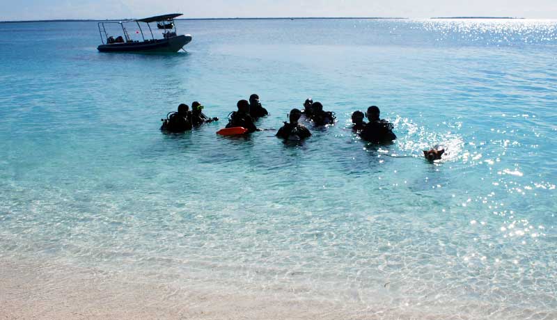 Scuba Do Zanzibar crew in the shallow water on scuba in front of the dive centre in crystal clear water with boat in background